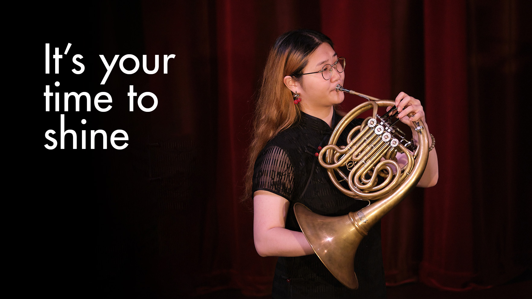 "It's your time to shine". A female Asian student playing the French horn, surrounded with by red curtains on stage.
